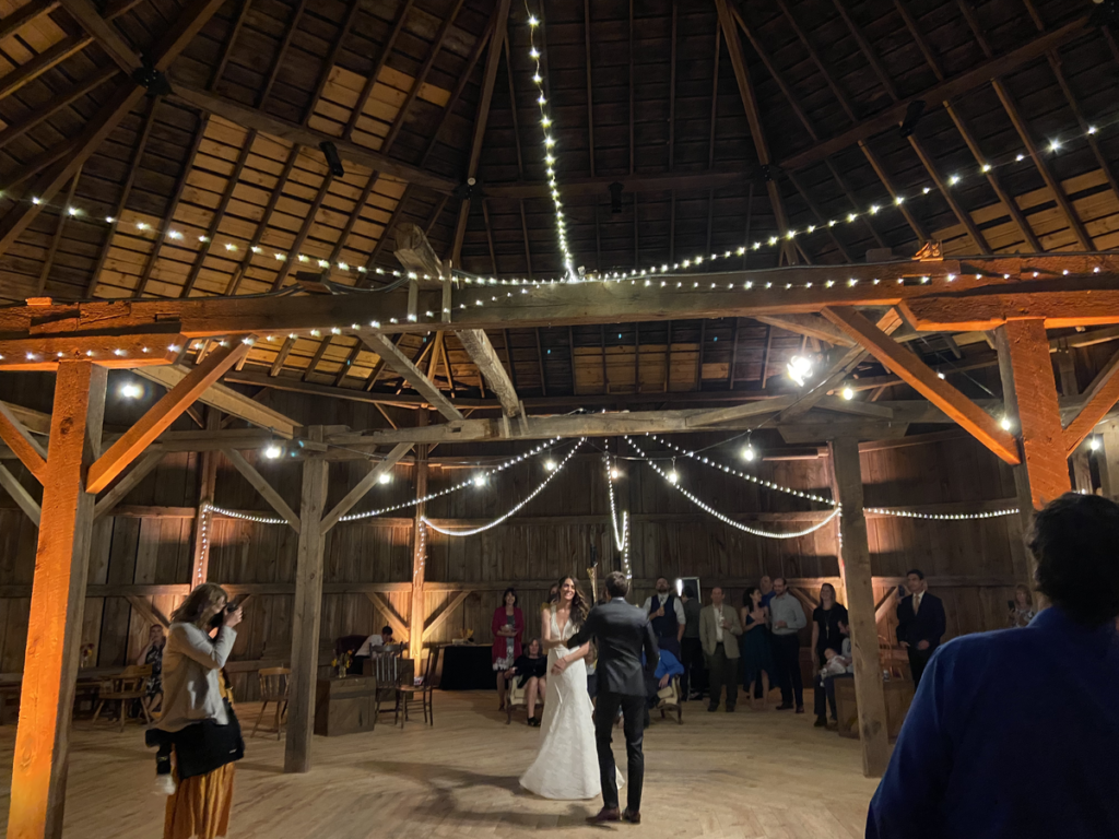 Bride and groom dancing in barn with woman photographing and white string lights above